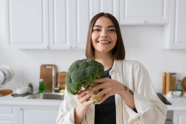 Позитивная молодая женщина смотрит в камеру, держа свежий брокколи на кухне — стоковое фото
