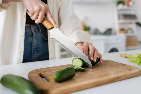 Cropped view of woman cutting ripe cucumber on chopping board in kitchen — Stock Photo