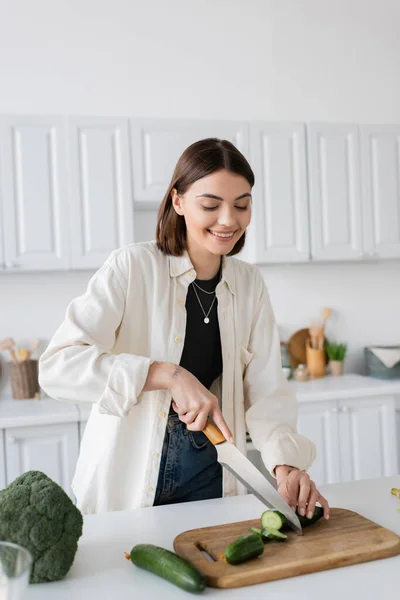 Cheerful tattooed woman cutting cucumber near broccoli and bowl in kitchen — Stock Photo