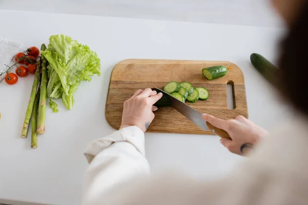 Вид сверху татуированной женщины, режущей огурец рядом с овощами на кухне — стоковое фото