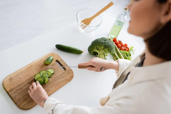 Cropped view of blurred woman cutting cucumber near fresh vegetables in kitchen — Stock Photo