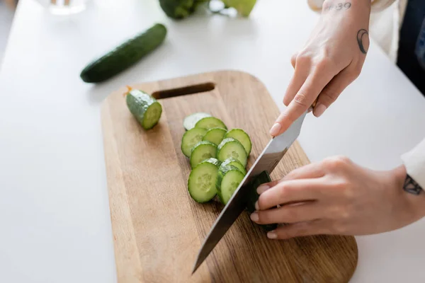 Vue recadrée d'une femme tatouée coupant du concombre à bord dans la cuisine — Photo de stock