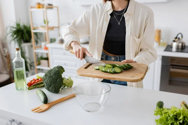 Vista ritagliata della donna che tiene il tagliere con cetrioli tagliati vicino a ciotola e verdure in cucina — Foto stock