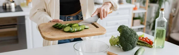 Обрізаний вид жінки, що тримає розрізаний огірок на обробній дошці біля миски на кухні, банер — стокове фото