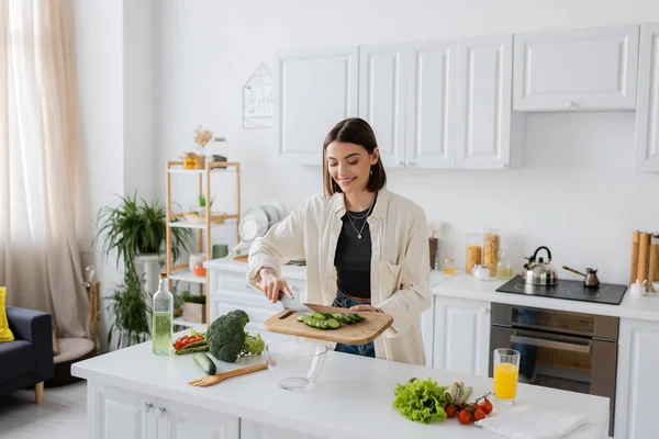 Mulher alegre segurando pepino cortado na tábua de corte enquanto cozinha salada na cozinha — Fotografia de Stock