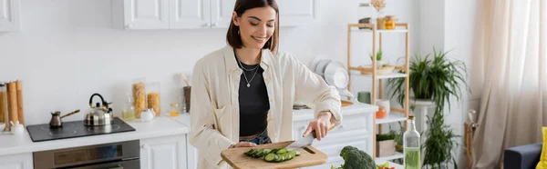 Cheerful woman holding sliced cucumber on chopping board in kitchen, banner — Stock Photo