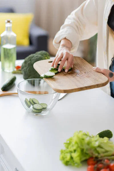 Ausgeschnittene Ansicht tätowierter Frau, die Gurkenscheiben in Schüssel in der Küche hinzufügt — Stockfoto