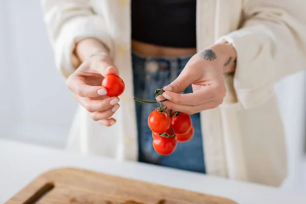 Vue recadrée d'une femme floue tenant des tomates cerises mûres près de la planche à découper dans la cuisine — Photo de stock
