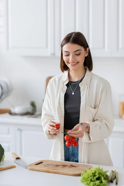 Positive Frau hält Kirschtomaten neben Gemüse und Schneidebrett in der Küche — Stockfoto