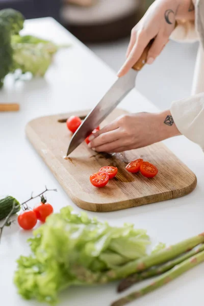 Cropped view of tattooed woman cutting cherry tomatoes near vegetables in kitchen — Stock Photo