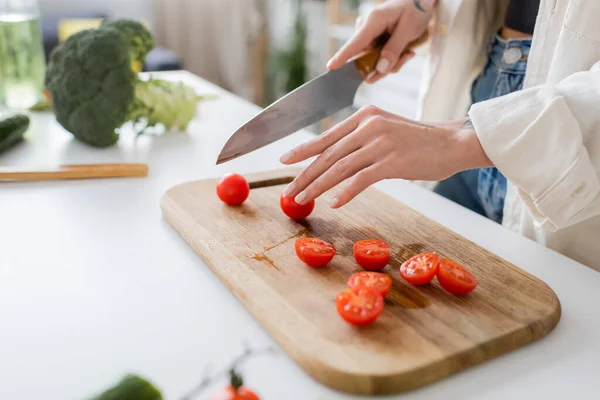 Cropped view of young woman cutting cherry tomatoes on chopping board in kitchen — Stock Photo