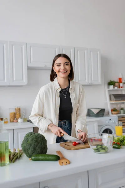 Fröhliche junge Frau schneidet Kirschtomaten neben Gemüse und Orangensaft in der Küche — Stockfoto