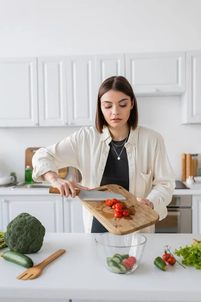 Mulher morena derramando tomates cereja na tigela enquanto cozinha salada na cozinha — Fotografia de Stock
