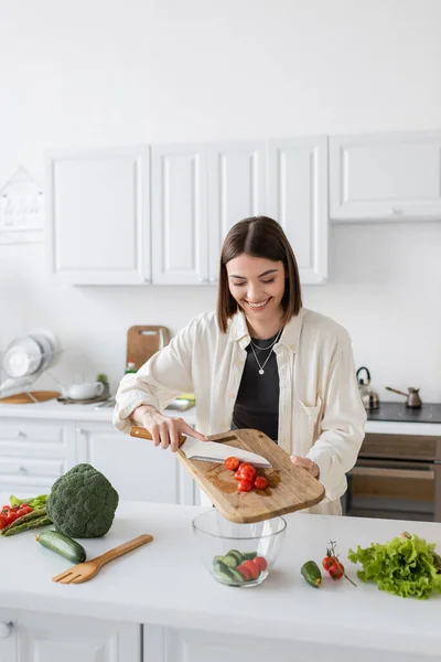 Mujer positiva vertiendo tomates cherry en un tazón mientras cocina ensalada fresca en la cocina - foto de stock