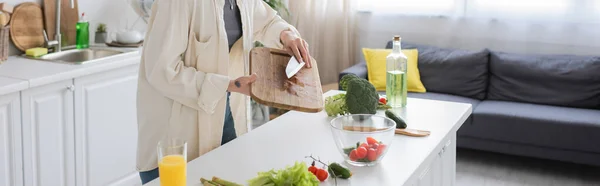 Vista recortada de mujer tatuada sosteniendo cuchillo y tabla de cortar mientras cocina ensalada fresca en la cocina, pancarta - foto de stock
