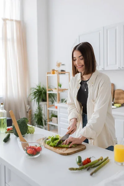 Позитивна брюнетка жінка ріже салат під час приготування салату на кухні — стокове фото