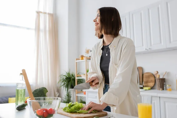 Усміхнена молода жінка розрізає салат біля апельсинового соку та салату на кухні — стокове фото