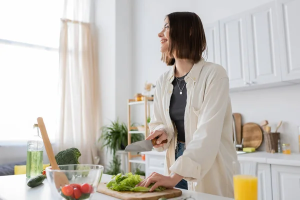 Sorridente giovane donna taglio lattuga vicino succo d'arancia e insalata in cucina — Foto stock