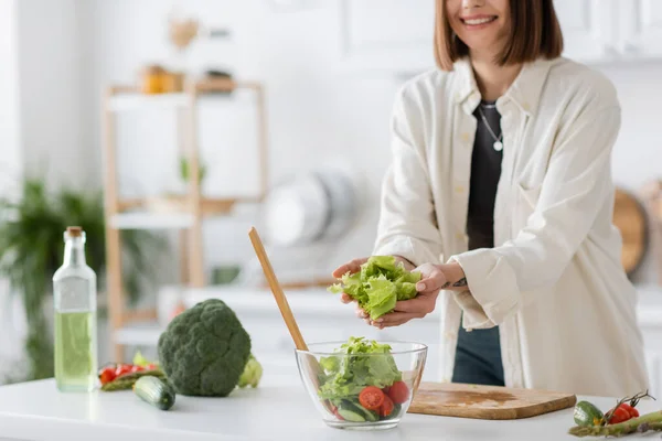Cropped view of smiling woman putting lettuce in bowl with salad in kitchen — Stock Photo