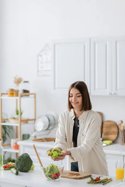 Mulher morena alegre colocando alface na tigela com salada na cozinha — Fotografia de Stock