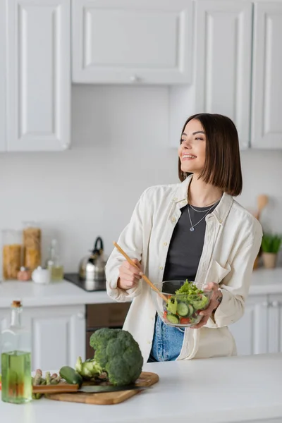 Улыбающаяся женщина держит миску с салатом рядом со свежими овощами на кухне — стоковое фото