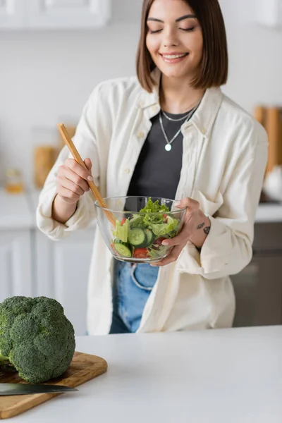 Jovem mulher positiva misturando salada fresca perto de brócolis na placa de corte na cozinha — Fotografia de Stock