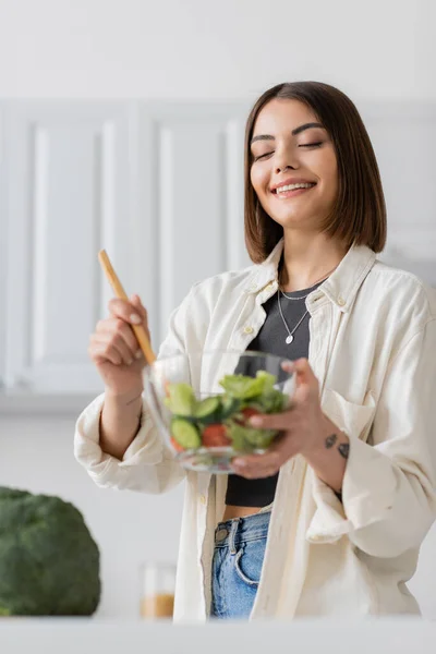 Cheerful tattooed woman mixing fresh salad in kitchen at home — Stock Photo