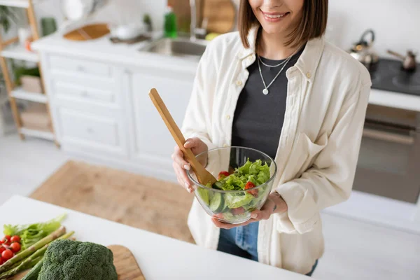 Обрезанный вид улыбающейся женщины, держащей свежий салат в миске на кухне — стоковое фото