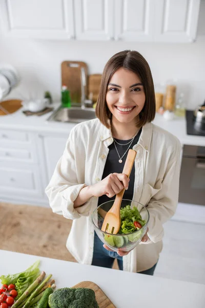 Jovem mulher positiva segurando salada fresca e olhando para a câmera na cozinha — Fotografia de Stock