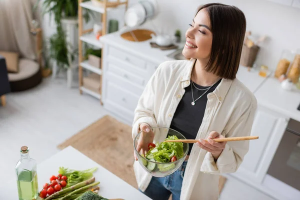 Беззаботная женщина держит свежий салат возле овощей и оливкового масла на кухне — стоковое фото