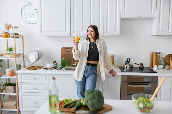 Smiling brunette woman holding orange juice near blurred salad in kitchen — Stock Photo