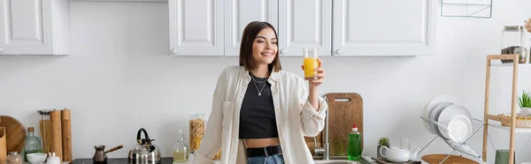 Femme brune positive tenant du jus d'orange dans la cuisine, bannière — Photo de stock