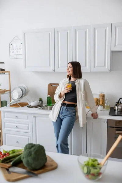 Donna spensierata che tiene il succo d'arancia vicino all'insalata sfocata sul piano di lavoro in cucina — Foto stock