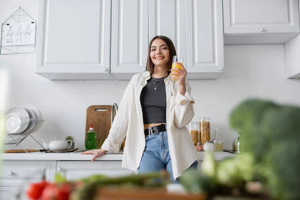 Positive woman holding orange juice near blurred vegetables in kitchen — Stock Photo