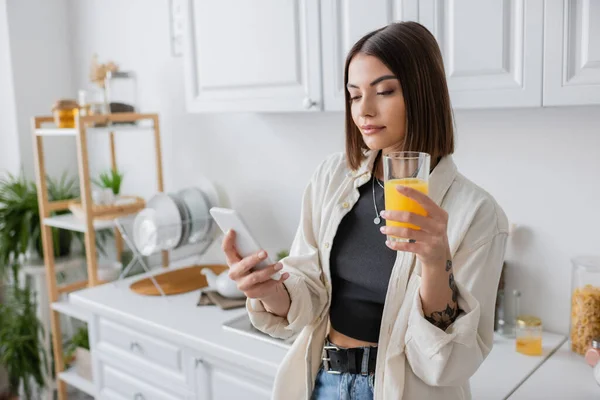 Mujer morena sosteniendo jugo de naranja y el uso de smartphone en la cocina - foto de stock