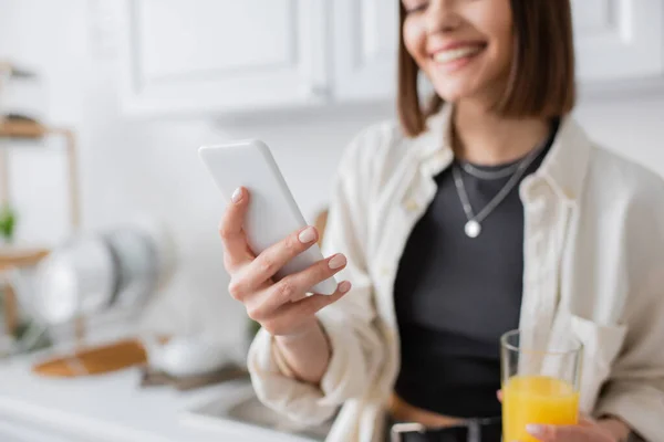 Vista recortada de una mujer sonriente sosteniendo jugo de naranja y usando el teléfono celular en la cocina - foto de stock