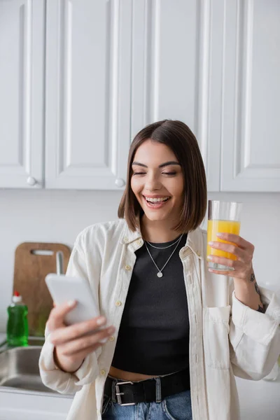 Mulher morena alegre segurando suco de laranja e usando smartphone desfocado na cozinha — Fotografia de Stock