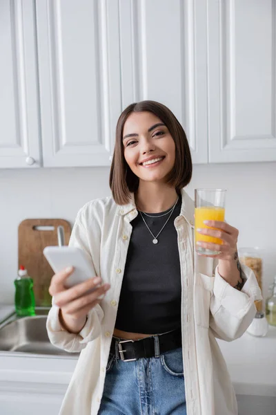 Mujer joven sonriente sosteniendo jugo de naranja y teléfono inteligente mientras mira a la cámara en la cocina - foto de stock