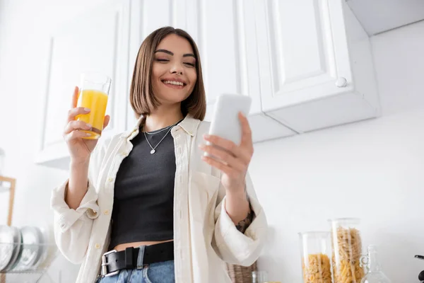 Low angle view of smiling woman using smartphone and holding orange juice in kitchen — Stock Photo