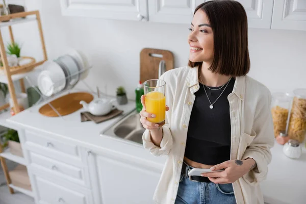 Веселая молодая женщина держит мобильный телефон и апельсиновый сок на кухне — стоковое фото