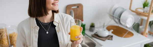 Vista cortada de mulher morena sorridente segurando suco de laranja fresco na cozinha, banner — Fotografia de Stock