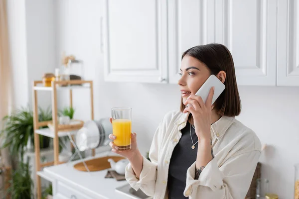 Молодая женщина разговаривает на смартфоне и держит апельсиновый сок на кухне — стоковое фото