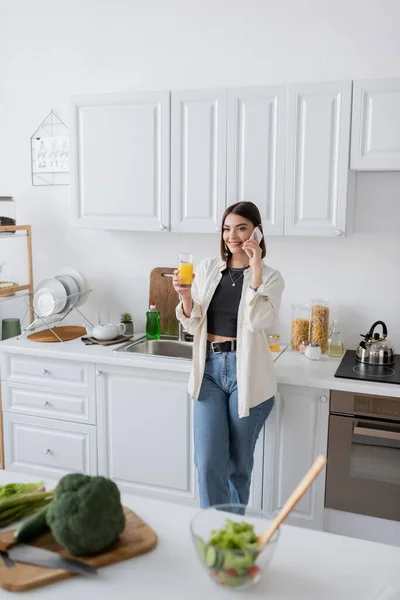 Cheerful young woman talking on cellphone and holding orange juice in kitchen — Stock Photo