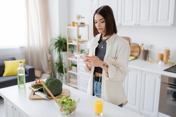 Femme tatouée utilisant un smartphone près du jus d'orange et de la salade fraîche dans la cuisine — Photo de stock