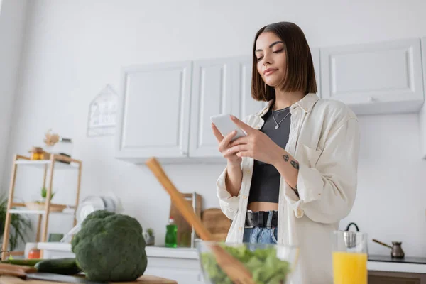 Vue à angle bas de la femme tatouée utilisant un téléphone portable près des légumes flous et du jus d'orange dans la cuisine — Photo de stock