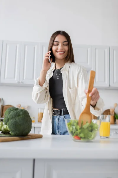 Mulher sorridente falando no smartphone e misturando salada fresca na cozinha — Fotografia de Stock