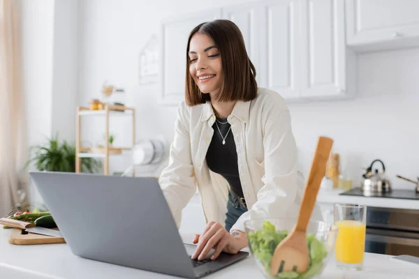 Femme brune joyeuse utilisant un ordinateur portable près de la salade et du jus d'orange dans la cuisine — Photo de stock