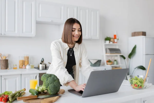Positive woman using laptop near ripe vegetables and orange juice in kitchen — Stock Photo