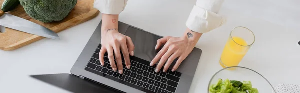 Cropped view of tattooed freelancer using laptop near orange juice and salad in kitchen, banner — Stock Photo