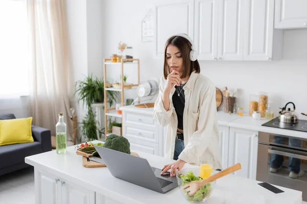 Young woman in headphones using laptop near salad and vegetables in kitchen — Stock Photo
